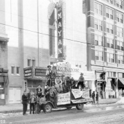 Men pose on a truck near the Mayan Theater at 110 Broadway in the Speer neighborhood of Denver, Colorado. The men wear cowboys hats and chaps. Signs read: "Warm Air Furnace Heating Tin and Galvanized Iron Work," "Wheeler Organist," "Gala Opening Fox Mayan Theatre Tonight," "Are Always Welcome at the Park Lane Hotel," "Beauty Nook," and "South Broadway National Bank."