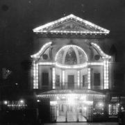Nighttime view of the Orpheum theater at 1537 Welton Street in downtown Denver, Colorado. The three-story building is illuminated with electric lights and has an ornate facade. A clock is on the roof.