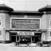 View of the Ogden Theater on Colfax Avenue in Denver, Colorado. The two-story brick building has a tile roof and ornate entryway. Automobiles and a bicycle are parked nearby. A sign reads: "This Theater Will Open Thurs. Sept. 6th."