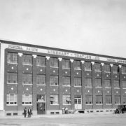 Men stand near the Minehart-Traylor Company building at the intersection of 25th (Twenty-fifth) Street, Broadway, and Walnut Street in the Five Points neighborhood of Denver, Colorado. The three-story brick building has decorative brickwork and contrasting trim. Lettering on the building reads: "Mintra Paints Minehart-Traylor Co."