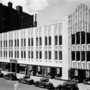 View of the art-deco style Paramount Theater building, designed by Temple Hoyne Buell, at 16th (Sixteenth) and Glenarm Streets in downtown Denver, Colorado. Automobiles are parked nearby. Signs in the window read: "Richard Barthelmess Dawn Patrol Tabor Theater [...]" and "New Home Colorado Chiropractic University."