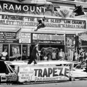 Men in suits perform stunts on a trampoline near the Paramount Theater designed by Temple Hoyne Buell at 16th (Sixteenth) and Glenarm Streets in downtown Denver, Colorado. A woman in costume and spectators stand nearby. Signs on the building read: "Paramount," "Basil Rathbone 'The Black Sleep' Lon Chaney [...]an Donlevy 'The Creeping Unknown' Margia Dean," "Akim Tamiroff, John Carradine, Bela Lugosi," "All New Double Shock S Horror Show," "It's Cool Inside," and "Look Gave 'Trapeze' a 6 Page Story."