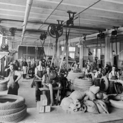 Interior view of the Gates Rubber Company in Denver, Colorado; shows men working with belt-driven machinery and rubber tires.