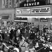 People crowd the entrance of the Denver Theater at 1545 Glenarm Place in Denver, Colorado; band members wear helmets, and the neon marquee reads: "Kit Carson," and "Charlie Chan at the Wax Museum."