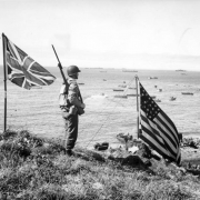 Jim "Wyoming" Penton, a member of the 10th Mountain Division 87th Regiment Service Company (SVC), stands on a bluff over Broad Beach on Kiska Island in Alaska; U.S. and British flags are nearby. The 87th Mountain Infantry Regiment was in the Aleutians as a part of Amphibious Task Force 9. Boats and amphibious vehicles are in the water in the distance.