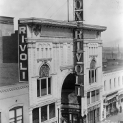Rooftop view of the Rivoli Theater at 1741 Curtis Street in downtown Denver, Colorado. The four-story building has an ornate entryway decorated with electric lights. Signs read: "Fox Rivoli," "Thos. H Ince's Great Drama 'The Cup of Life,'" and "Hotel Elwood."