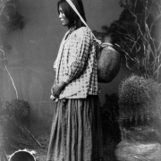 Standing studio portrait vignette of a Native American San Carlos Apache woman posed in profile carrying a pitch covered basket jar with a head strap and wearing patterned blouse and skirt; props include baskets, cactus and brush.