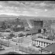 View of Denver, Colorado, shows downtown buildings including the Arapahoe County Courthouse. Clouds top front range mountains; signs read: "I.A. (Mrs. Ida Alkire) Lehman Specialty Shop (or The Toggler Shop, 1538 California)", "Overland Cars," and "YMCA."