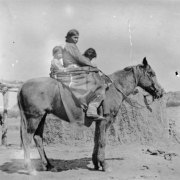 A Native American (Navajo) woman and children pose on horseback near an earthen hogan.