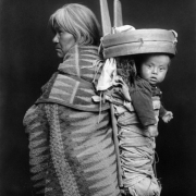 A Native American (Navajo) woman poses with a child in a cradleboard on her back. The woman wears a blanket.
