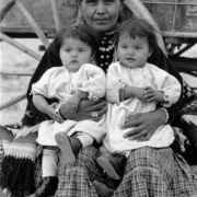 A Native American (Navajo) woman poses outdoors with her twin children. They sit near a wagon.