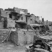 A Native American (Hopi) woman stands near a group of adobe cluster homes at Oraibi Pueblo, Arizona. The homes have chimney pots and wooden vigas.