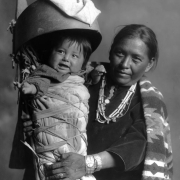 Studio portrait (sitting) of a Native American (Navajo) mother and child. The baby is in a cradleboard. The mother wears necklaces and silver bracelets.