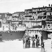 Native American (Pueblo, Zuni) children and women and white men and women stand on and near adobe and stone cluster homes at Zuni Pueblo, New Mexico. The buildings have chimney pots, vigas, and wooden ladders.