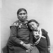 Sitting studio portrait of Beautiful Hill, a Native American (Plains) woman, and Village Maker, a Omaha boy. The woman wears a dark dress with shoulder conchos, bead necklaces, a choker, and earrings. The child, standing next to her, wears leggings, a shirt, and bead and shell necklaces.
