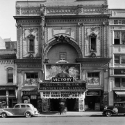 View of the Victory Theater at 1620 Curtis Street in Denver, Colorado; shows the arched entry, entablature, and cars. Marquee and neon signs read: "Here Comes the Band, Wallace Ford, Another Face," and "Sobule's Liquors."