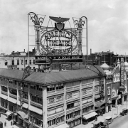 View of the America theater at 16th (Sixteenth) and Curtis Streets in Denver, Colorado. Cars and people are in the streets; electric roof sign depicts an eagle and shield and reads: "America Theater - See America First - Nazimova In A Dolls House." The Empress Theater is next door.