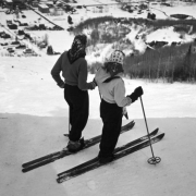 Two women skiers in Aspen, Colorado stand at the top of the ski hill facing downwards. Both women wear trousers, light jackets and scarves on their head.  Trees are on either side of the ski slope, and the town is at the base of the hill.
