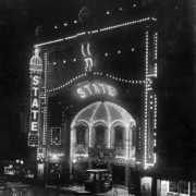 Night view of the State Theater in Denver, Colorado; electric lights ornament the arch, facade, and sign. Showcards read: "My Own Pal - Tom Mix."