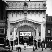 View of the Strand theater in Denver, Colorado; shows an ornate entry with entablature and electric lights. People, bicycles and cars are in the street; marquee reads: "Frank Mayo in Dr. Jim," signs read: "Dr. Murphy's Root Beer," "Shoe Shine," and "Razor Blade Hospital."