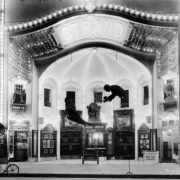 Night view of the Strand theater in Denver, Colorado; shows an ornate entry with entablature and electric lights. Display depicts a spider web. Marquee reads: "Alice Joyce - The Prey" and "Bride 13 the Torture Chamber." Signs read: "Dr. Murphy's Root Beer" and "Shoe Shine."
