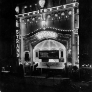 Night view of the Strand theater in Denver, Colorado; shows an ornate entry with entablature and electric lights. Display depicts a false front. Marquee reads: "Sunset Jones." Signs read: "Dr. Murphy's Root Beer" and "Shoe Shine."