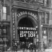 View of the Tabor theater 16th (Sixteenth) Street entrance in Denver, Colorado. People, boys, and bicycles are by the arched entry and ticket window; electric signs read: "Continuous Vaudeville 5 [cents] 10 [cents]." Poster advertises "The Balkan Princess."