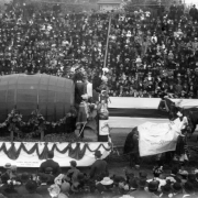 Men ride on a horse-drawn float, part of the Festival of Mountain and Plain, in a parade in Denver, Colorado. The float depicts a king with a sword and a keg of beer. Men on the float wear costumes that include hats decorated with feathers, capes, and swords. The horses wear decorative cloth shrouds with lettering that reads: "Neef Bros Brewing Co." Spectators sit in grandstands nearby.