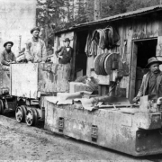 View of men in chain of ore carts being pulled by General Electric engine with frame building, a man stands beside the door, Colorado.