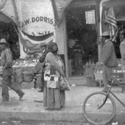 A Native American (Maricopa) woman stands on the sidewalk near a market on Washington Street in Phoenix, Arizona. She holds a basket and wears a blanket. A sign reads: "J. W. Dorris [W]ash". Shows a Native American man, white men, and a bicycle.