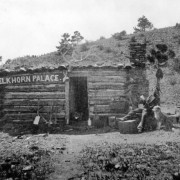 A man sits with his dog outside of a hewn log cabin near Rosita (Custer County), Colorado. The cabin has a single doorway, a rack of elk horns over the door, a chimney, and a sign reading: "Elkhorn Palace."