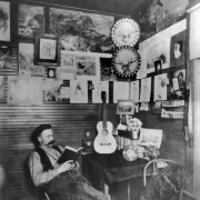 A miner relaxes with a book in his cabin, a dog sits beside him on a chair, at Camp Bird mine, Ouray County, Colorado. A guitar, books, plates, and flowers are on the table in front of him; pinup girls, and nature scenes hang on the walls.