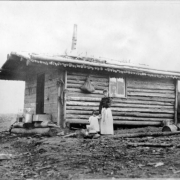 A woman and a young girl pose near a log cabin on a snow dusted landscape as a man driving a wagon approaches in the background, Colorado. The cabin has a plank front with timber walls and roof.