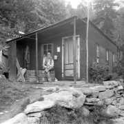 A miner poses outdoors on a trunk on the porch of his cabin probably in Colorado. He wears a cap with a miner's light.