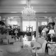 Interior view of the Claude K. Boettcher residence, in Denver, Colorado; decor includes upholstered couches, chairs, ottomans, palms, a sculpture of a woman, lamps, a crystal chandelier, classical columns, and a planter with bas-relief ornaments.