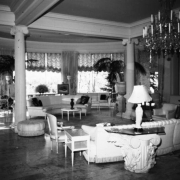 Interior view at the Claude K. Boettcher residence, in Denver, Colorado; living room decor includes curved couches, chairs, an ottoman, glass tables, Corinthian capitols, a crystal chandelier, palms, and curtains.