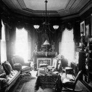 Interior view of the F. J. Bancroft house in Denver, Colorado; decor includes Oriental rugs, a hanging lamp, a taxidermied heron, ewers, a Buddha, framed art, and upholstered chairs.