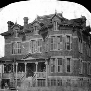 View of an orphanage in Denver, Colorado; the brick residence has gables, widow's walks, and bays. People pose on the covered porch.