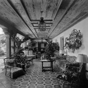 View of a covered patio in Denver, Colorado. Coffered ceilings, wrought iron lamps, couches, chairs, potted plants, and a tile floor comprise the decor. An open plaza is to the side.