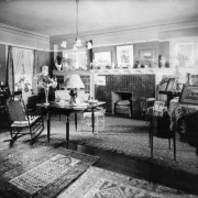 Interior view of a house in Denver, Colorado; decor includes Oriental rugs, rocking chairs, a fireplace, lamps, flower vases, porcelain, photos, and a piano.