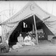 Portrait of women, girls, and dogs in a tent in (probably) Denver, Colorado; one plays a mandolin. A star / crescent motif is on the striped canvas; embroidery and quilts are inside.