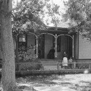 Boys, women, and a girl pose outdoors near the John L. Dailey house at 4th (Fourth) Avenue and Broadway in Denver, Colorado. The wood frame house has a covered porch and is surrounded by a metal fence.