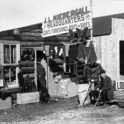 Two men and a dog are outside a log store for miners in Fremont (Cripple Creek, Teller County), Colorado. The sign above the store reads "J.L. Niebergall, Headquarters for Gents Furnishings Boots & Shoes."  Shirts, pants, jackets, gloves, socks, shoes and boots are arranged on boxes and hung on beams outside the cabin.  A sign outside the cabin includes price list. Suspenders hang in the window. A sign: "Lodging," is on a stack of building materials beside a plank building under construction.