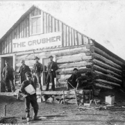 Six men are gathered on the front deck of the The Crusher, the first newspaper in Fremont (Cripple Creek, Teller County), Colorado.  Oakley Spell, a young boy, holds a bundle of newspapers under his arm; a sign on the log building reads: "The Crusher."