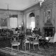 Interior view of the living room in the Lawrence Cowle Phipps house at 3400 Belcaro Drive in the Belcaro neighborhood of Denver, Colorado. Chairs, tables, a sofa, fireplace, a chandelier, and portraits comprise decor.