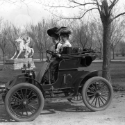 Women ride in an early automobile in City Park, Denver, Colorado. They wear dresses and hats decorated with feathers. A statue, 'The Cowboy,' created by Alexander Phimister (A. P.) Proctor, is in the distance.