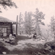 A prospector and guide, with a pipe and a dog and puppies, sits by his log cabin in Teller County, Colorado. The cabin is built into a hill and has a sod roof. A cast-iron wood stove is outside the cabin, a sign reads "Burros for hire." Shows a sawhorse and a can with a spout.