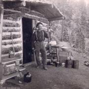 A prospector with a sledgehammer stands outside his chink and log cabin in Teller County, Colorado; shows a cast-iron stove outside the door of the cabin, a jug, broom, lunch pail, pans, cans, and a dog and her puppies under a table.