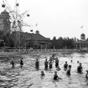 Men, women and children swimmers wade in Lake Rhoda at Lakeside Amusement Park in Lakeside (Jefferson County), Colorado. A lifeguard sits in a row boat and several people lounge on the beach. The Staride ferris wheel, a pavilion and the Casino Tower entrance are in the background.