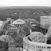 Cars speed along the tracks of the roller coaster at Lakeside Amusement Park in Lakeside (Jefferson County), Colorado. A large arch with a sign reading: "Welcome" spans the sidewalk leading to a large ballroom building. People stroll along the walkways. Lake Rhoda is in the distance.